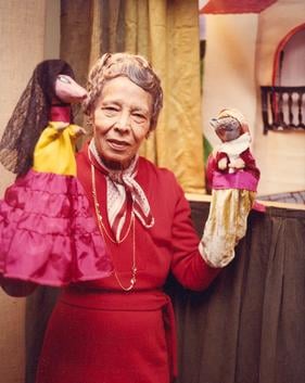A full color photo of Pura Belpré standing wearing a red dress and scarf with a puppet on each hand in front of a stage with a house and a curtain. Photo Source: Wikipedia